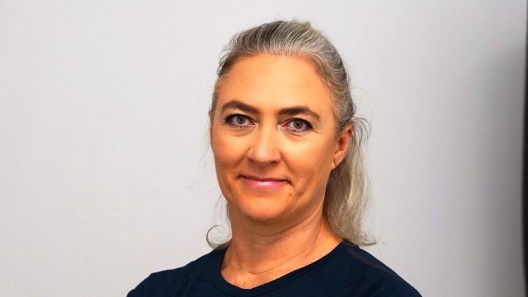 Frau mit grauen langen Haaren und lächelndem Gesicht vor neutralem Hintergrund.